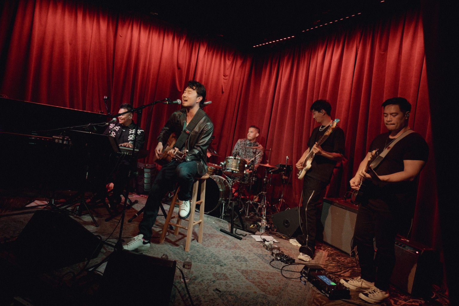 Kevin Michael Chong performing behind Simu Liu and the band on the red-curtain stage at Hotel Café